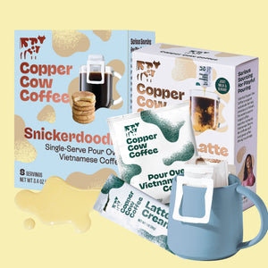 Copper Cow Coffee Snickerdoodle Vietnamese Coffee Kit shown with two product boxes, several single-serve packets, a blue pour-over mug, and a small puddle of creamer on a pale yellow background.